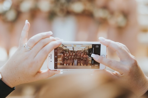 A person is taking a photo with a smartphone, capturing a group of people posing in a well-lit setting. The focus is on the smartphone screen displaying the group image. The setting suggests a social gathering or event.