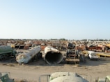 A large scrapyard filled with rusted and abandoned vehicles and machinery, including tankers, trucks, and other heavy equipment spread across a dusty, barren landscape.