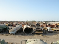 A large scrapyard filled with rusted and abandoned vehicles and machinery, including tankers, trucks, and other heavy equipment spread across a dusty, barren landscape.