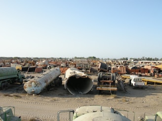 A large scrapyard filled with rusted and abandoned vehicles and machinery, including tankers, trucks, and other heavy equipment spread across a dusty, barren landscape.