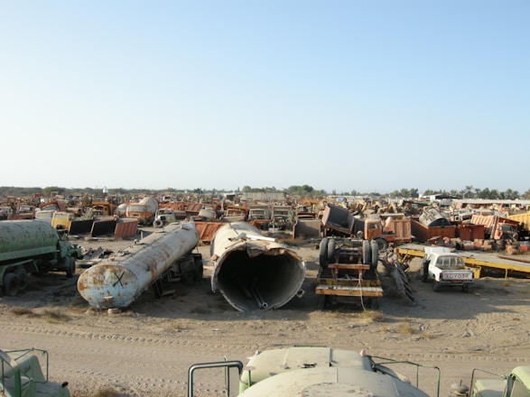 A large scrapyard filled with rusted and abandoned vehicles and machinery, including tankers, trucks, and other heavy equipment spread across a dusty, barren landscape.