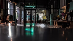 A modern cafe with glass doors and large windows, featuring a neon sign at the top. Inside, there are several tables and chairs, with coats hanging on a rack to the left. The interior is dimly lit, with reflections visible on the polished surface of a counter in the foreground.