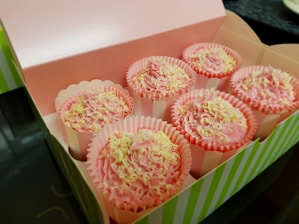 A box containing six cupcakes with pink frosting and white chocolate shavings. The box is open and has a pink interior with green and white striped edges. Each cupcake is in its own paper liner, which is pink and has a scalloped edge.