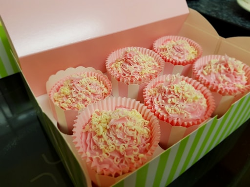 A box containing six cupcakes with pink frosting and white chocolate shavings. The box is open and has a pink interior with green and white striped edges. Each cupcake is in its own paper liner, which is pink and has a scalloped edge.