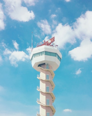 A tall control tower with a modern design stands against a bright blue sky with scattered white clouds. The tower features a windowed observation area at the top and an exterior staircase spiraling down its length. Antennas and equipment are visible on the top structure.