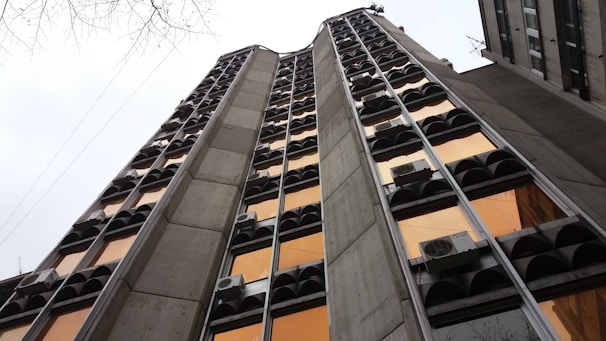 A tall, narrow building with an architectural design featuring multiple vertical concrete columns and rows of windows with reflective orange glass. The building facade includes air conditioning units attached below some windows. The sky is overcast, and bare branches of a tree can be seen in the top left corner.