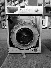 An old, dismantled washing machine is placed on a city sidewalk. The drum is exposed, and various components are missing or broken. Surrounding it are urban elements like apartment buildings and parked cars.