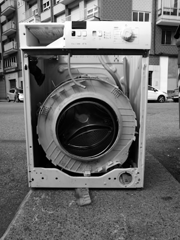 An old, dismantled washing machine is placed on a city sidewalk. The drum is exposed, and various components are missing or broken. Surrounding it are urban elements like apartment buildings and parked cars.