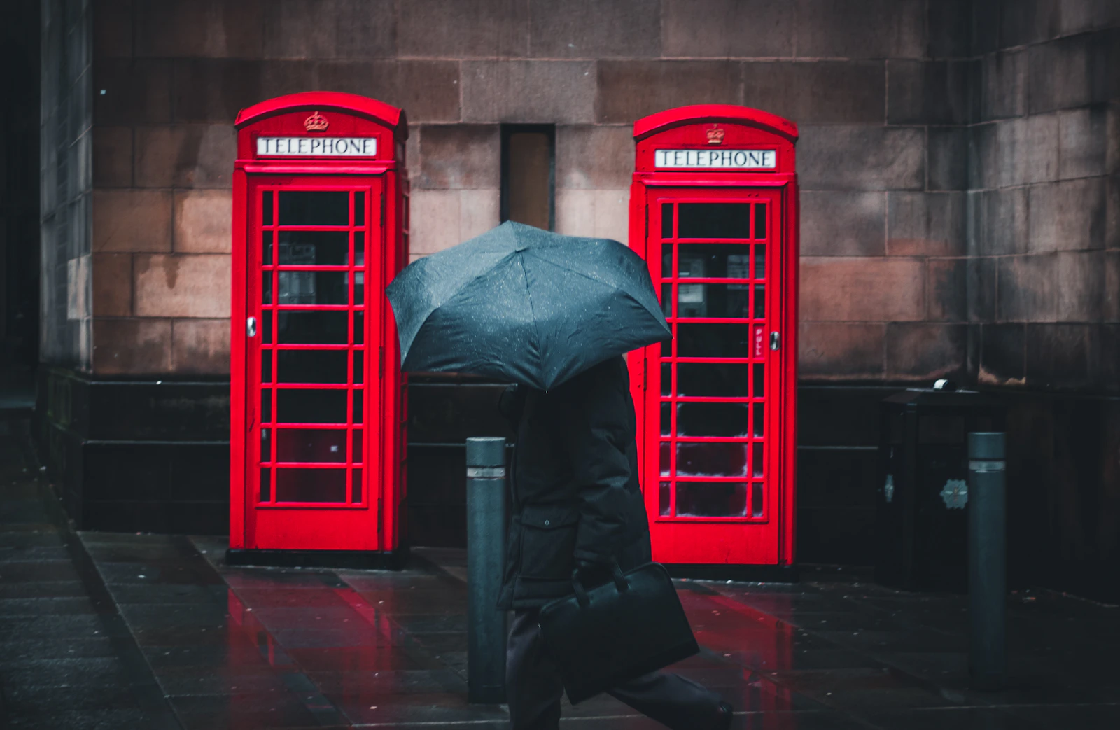 A person walking under an umbrella past red telephone boxes in the rain