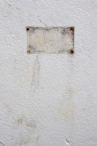 A close-up of a rough, textured white wall with a small, rectangular metal plate attached at the center. The plate is secured by four rusty screws, one at each corner. The metal appears weathered and slightly discolored, with faint hints of red and gray.
