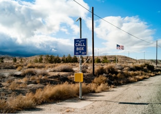 A roadside scene in a desert setting with a call box next to a gravel road. An American flag flutters in the distance, attached to a thin pole. The landscape features dry vegetation and rolling hills under a partly cloudy sky.