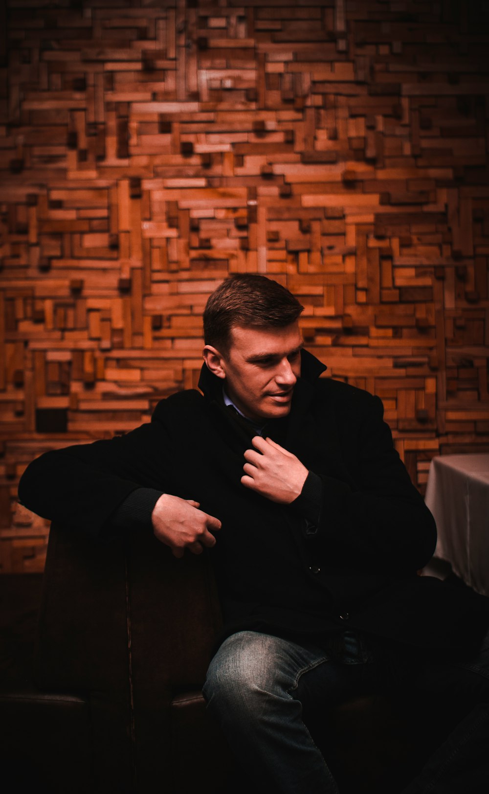 man in black coat and blue denim jeans sitting near brown wall
