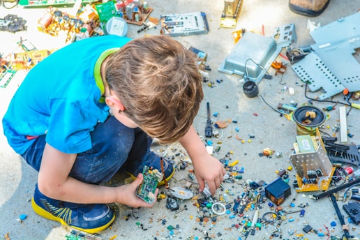 A child in a blue shirt and dark pants squats on the ground, surrounded by scattered electronic components. The child is examining a circuit board, showing a keen interest in the various parts. Nearby, there are various small and large electronic parts and debris.