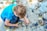A child in a blue shirt and dark pants squats on the ground, surrounded by scattered electronic components. The child is examining a circuit board, showing a keen interest in the various parts. Nearby, there are various small and large electronic parts and debris.
