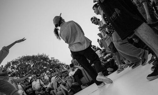 A lively outdoor gathering with a crowd of people engaged around someone performing or speaking. The focus is on a person wearing a cap, surrounded by others who are sitting or standing, watching intently. The scene feels energetic with several individuals capturing the moment on cameras. Trees are visible in the background, enhancing the casual and relaxed atmosphere.