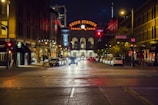 A city street at night, featuring Union Station with its illuminated neon sign reading 'Travel by Train' in the background. The street is lined with parked cars and buildings adorned with various signs and lights. The area is quiet and empty, with a warm glow from streetlights casting a cozy ambiance.