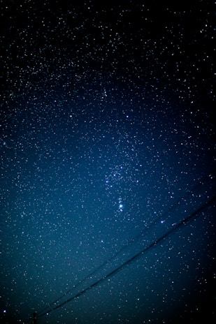 A star-filled night sky with a prominent constellation in the center. There are faint power lines crossing diagonally at the bottom. The sky is clear, allowing the multitude of stars to shine through.