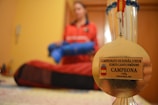 A gold medal is in focus against a blurred background of a person in a red outfit holding a blue object. The medal has an inscription for a junior karate championship, specifically for a female cadet kumite category. The background suggests an indoor setting.