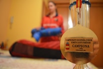 A gold medal is in focus against a blurred background of a person in a red outfit holding a blue object. The medal has an inscription for a junior karate championship, specifically for a female cadet kumite category. The background suggests an indoor setting.
