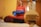 A gold medal is in focus against a blurred background of a person in a red outfit holding a blue object. The medal has an inscription for a junior karate championship, specifically for a female cadet kumite category. The background suggests an indoor setting.