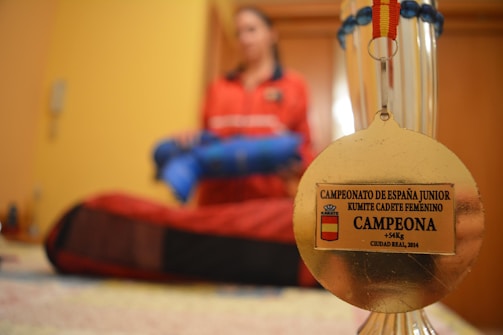 A gold medal is in focus against a blurred background of a person in a red outfit holding a blue object. The medal has an inscription for a junior karate championship, specifically for a female cadet kumite category. The background suggests an indoor setting.