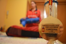 A gold medal is in focus against a blurred background of a person in a red outfit holding a blue object. The medal has an inscription for a junior karate championship, specifically for a female cadet kumite category. The background suggests an indoor setting.
