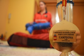 A gold medal is in focus against a blurred background of a person in a red outfit holding a blue object. The medal has an inscription for a junior karate championship, specifically for a female cadet kumite category. The background suggests an indoor setting.