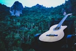 A classical guitar rests on a chair set against a lush, dense forest with prominent rock formations in the background. The scene is serene, highlighting the interaction between music and nature.