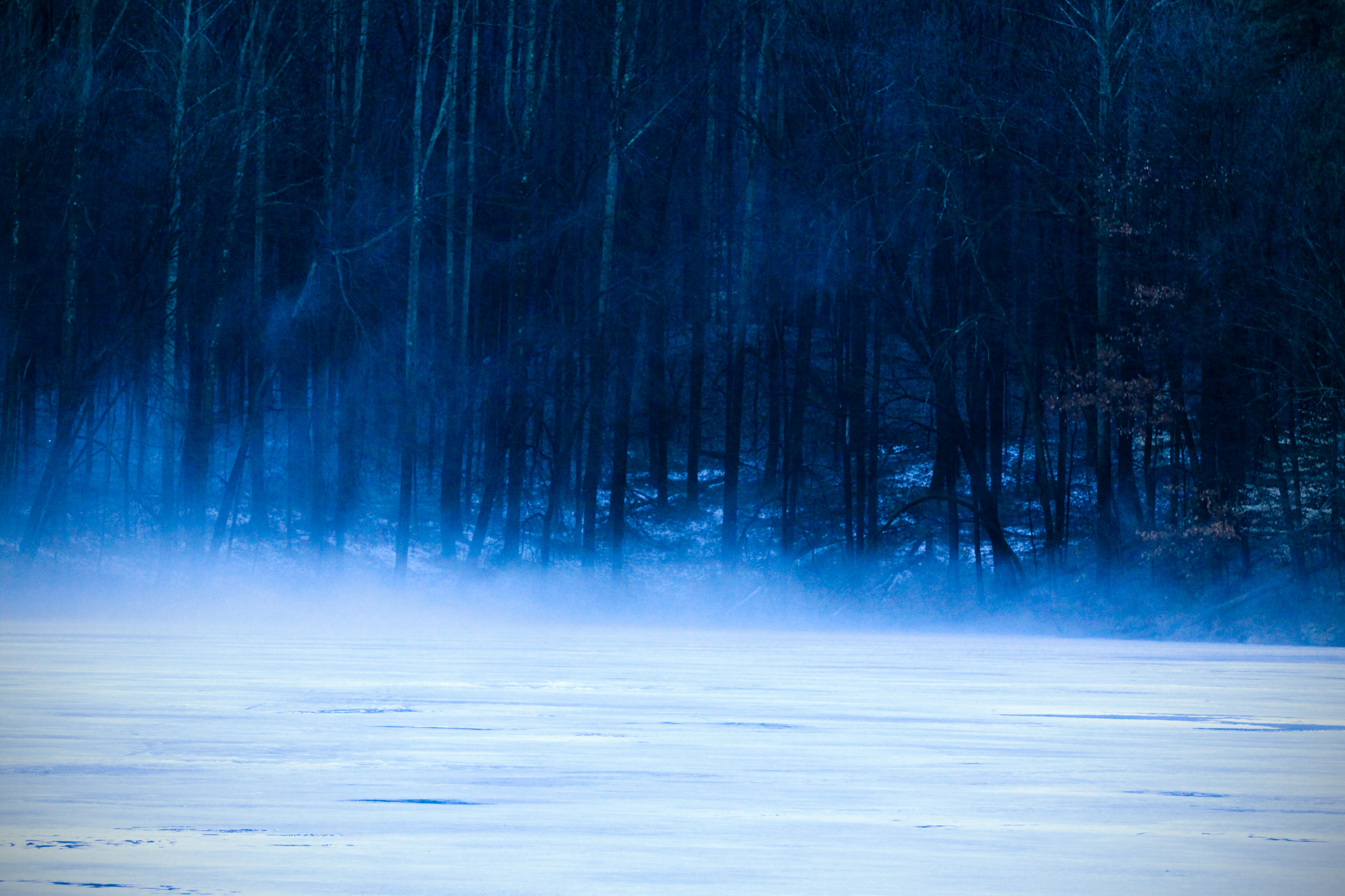 En frossen elv i forgrunnen med damp som stige opp. En mørk skog i bakgrunnen.