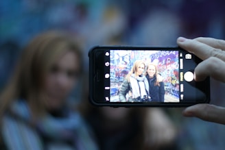 A close-up of a smartphone capturing a photo of two women standing in front of a graffiti-covered wall. The phone is held by a hand on the right side of the image, and the screen clearly shows the women smiling at the camera.