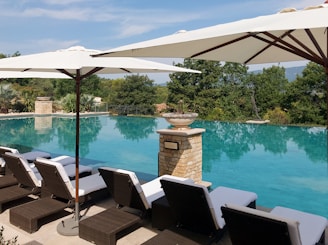 A serene outdoor pool area features multiple sun loungers with white cushions arranged neatly under large white umbrellas. The clear blue water of the infinity pool extends towards lush greenery, creating a tranquil and luxurious atmosphere. A decorative stone column with a planter adds elegance to the setting.
