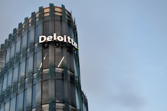 A modern glass building with sleek architectural design. The structure features a prominently displayed sign with the name 'Deloitte' in large, white letters. The facade consists of reflective glass panels, adding a contemporary aesthetic to the building. The sky in the background is clear and blue, enhancing the overall urban setting.