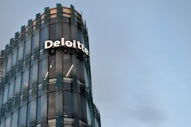 A modern glass building with sleek architectural design. The structure features a prominently displayed sign with the name 'Deloitte' in large, white letters. The facade consists of reflective glass panels, adding a contemporary aesthetic to the building. The sky in the background is clear and blue, enhancing the overall urban setting.