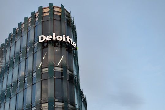 A modern glass building with sleek architectural design. The structure features a prominently displayed sign with the name 'Deloitte' in large, white letters. The facade consists of reflective glass panels, adding a contemporary aesthetic to the building. The sky in the background is clear and blue, enhancing the overall urban setting.