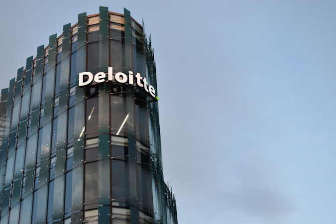 A modern glass building with sleek architectural design. The structure features a prominently displayed sign with the name 'Deloitte' in large, white letters. The facade consists of reflective glass panels, adding a contemporary aesthetic to the building. The sky in the background is clear and blue, enhancing the overall urban setting.