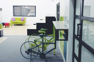 A modern indoor space with a combination of minimalist and colorful furnishings. Two bicycles, one black and one green, are parked near a wall-mounted desk. In the background, a green sofa with purple cushions is placed in front of a window with black bars. Several colorful stools are arranged near the sofa. There is a staircase with sharp geometric lines visible as well.