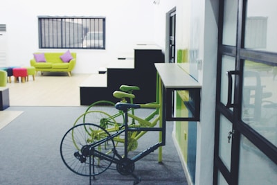 A modern indoor space with a combination of minimalist and colorful furnishings. Two bicycles, one black and one green, are parked near a wall-mounted desk. In the background, a green sofa with purple cushions is placed in front of a window with black bars. Several colorful stools are arranged near the sofa. There is a staircase with sharp geometric lines visible as well.