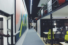 A modern office space with a long corridor featuring colorful wall art. On the right, there is an open-area workspace with desks and ergonomic chairs, some in bright green. The office is divided into two levels with potted plants visible on the upper level. A person is slightly visible on the far left, holding a door open.