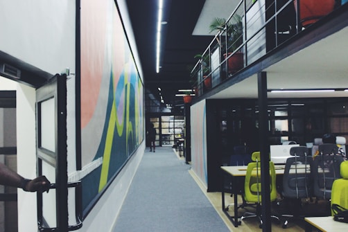 A modern office space with a long corridor featuring colorful wall art. On the right, there is an open-area workspace with desks and ergonomic chairs, some in bright green. The office is divided into two levels with potted plants visible on the upper level. A person is slightly visible on the far left, holding a door open.