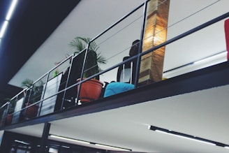 A modern indoor space with a metal railing and a row of potted plants including tropical palms. There is a tall, woven lamp providing warm light, and a blue cushion can be seen on a seat or couch. The ceiling has linear light features.