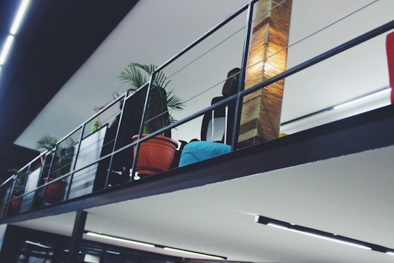 A modern indoor space with a metal railing and a row of potted plants including tropical palms. There is a tall, woven lamp providing warm light, and a blue cushion can be seen on a seat or couch. The ceiling has linear light features.