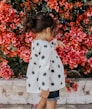 girl in black and white polka-dot long-sleeved blouse and blue shorts standing beside red petaled flowers during daytime