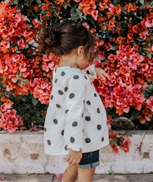 girl in black and white polka-dot long-sleeved blouse and blue shorts standing beside red petaled flowers during daytime