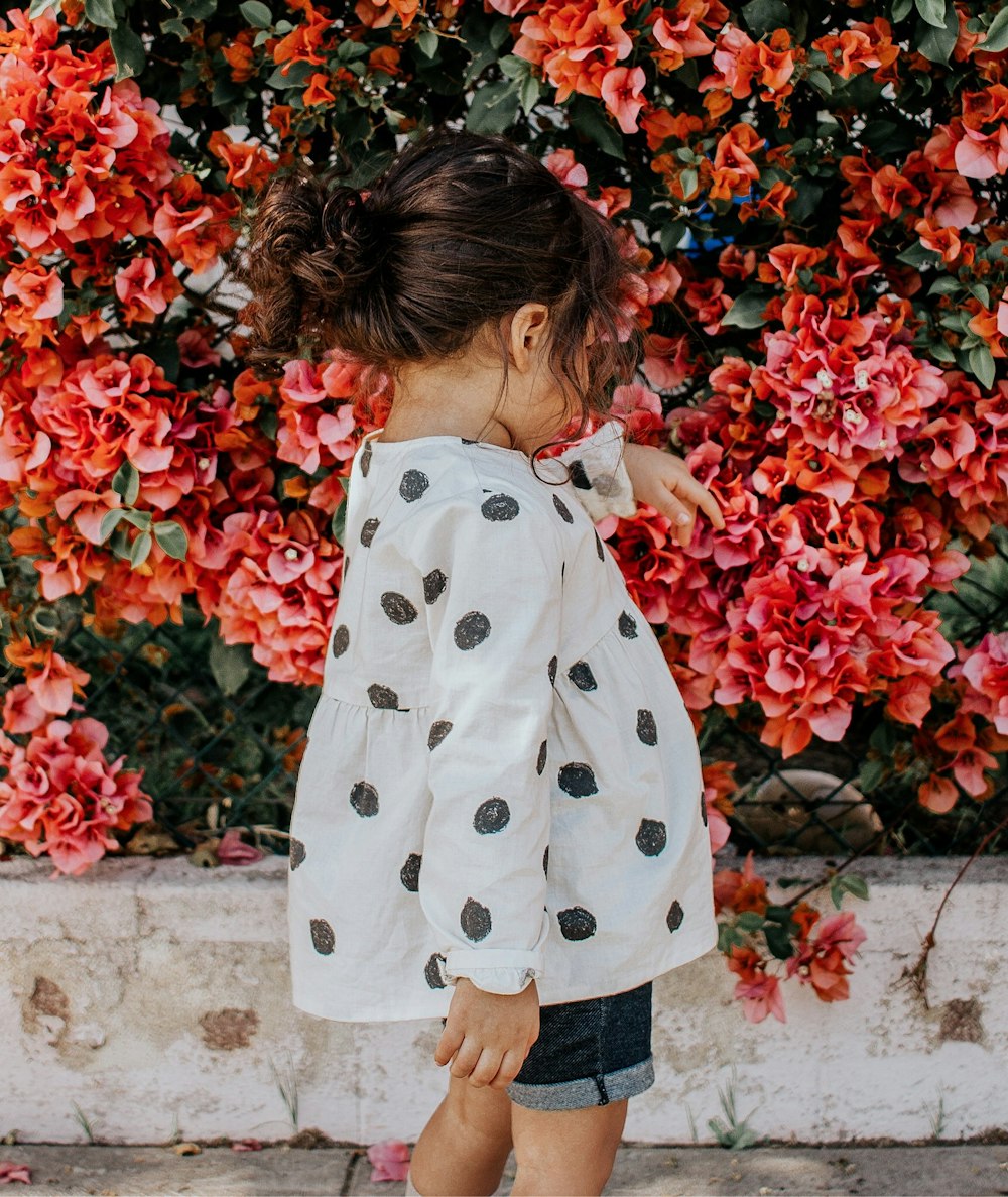 girl in black and white polka-dot long-sleeved blouse and blue shorts standing beside red petaled flowers during daytime