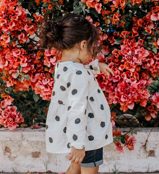 girl in black and white polka-dot long-sleeved blouse and blue shorts standing beside red petaled flowers during daytime