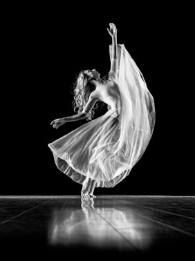 A graceful dancer is captured mid-movement, with one arm extended upward and the other out to the side. She wears a flowing, translucent skirt that swirls around her, creating a dynamic and ethereal effect. Her long hair cascades down her back as she balances elegantly on the wooden floor. The high contrast of the black background enhances the luminous quality of her dress and pose.