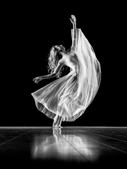 A graceful dancer is captured mid-movement, with one arm extended upward and the other out to the side. She wears a flowing, translucent skirt that swirls around her, creating a dynamic and ethereal effect. Her long hair cascades down her back as she balances elegantly on the wooden floor. The high contrast of the black background enhances the luminous quality of her dress and pose.