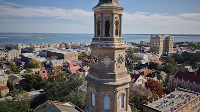 A tall stone clock tower stands prominently in a historic coastal cityscape, surrounded by densely packed buildings with colorful rooftops. In the background, a calm blue sea stretches under a slightly cloudy sky. The architecture suggests a blend of historical and modern influences.