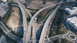 An aerial view of a complex highway interchange with several elevated roads crossing each other. Vehicles are seen traveling in both directions, surrounded by green spaces, urban buildings, and parking lots. Trees and vegetation line the roads, suggesting a mix of urban and natural environments.