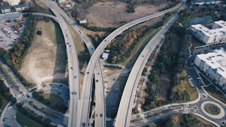 An aerial view of a complex highway interchange with several elevated roads crossing each other. Vehicles are seen traveling in both directions, surrounded by green spaces, urban buildings, and parking lots. Trees and vegetation line the roads, suggesting a mix of urban and natural environments.